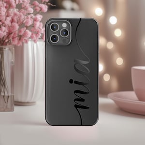 Könnte beinhalten: Schwarze Handyhülle mit dem Namen "mia" in Schreibschrift. Die Hülle hat ein geschwungenes Designelement auf der rechten Seite. Die Handyhülle liegt auf einer weißen Oberfläche mit einem verschwommenen Hintergrund mit rosa Blumen und einer rosa Tasse.