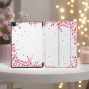 May include: A tablet case with a white interior and a black frame. The case is decorated with pink cherry blossom branches and falling petals. The case is open, revealing the interior and the design on the cover.