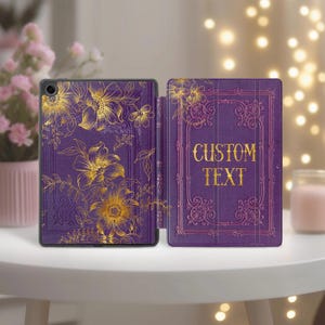 Könnte beinhalten: Eine lila Tablet-Hülle mit einem goldenen Blumendesign. Die Hülle ist geöffnet und zeigt das Innere mit den Worten "CUSTOM TEXT" in Gold. Das Design zeigt Blumen und Blätter. Die Hülle liegt auf einem weißen Tisch.