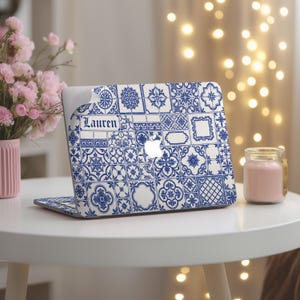 Puede incluir: Ordenador portátil con un diseño de azulejos azules y blancos. El portátil tiene el nombre "Lauren" impreso. Está sobre una mesa blanca con una vela rosa y un jarrón con flores rosas.