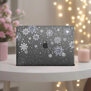 May include: A dark gray laptop with a winter-themed design. White snowflakes of various sizes are scattered across the surface. The Apple logo is visible in the center. The laptop is on a white table.