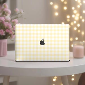 May include: A closed laptop with a yellow and white gingham pattern on the lid, featuring a black Apple logo. The laptop sits on a white table, with a vase of pink flowers and a candle in the background.