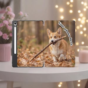May include: A black tablet case featuring a printed image of a tan and white Corgi dog holding a stick in its mouth while walking on fallen autumn leaves. The text "Your photo here" is printed in white on the right side of the case.