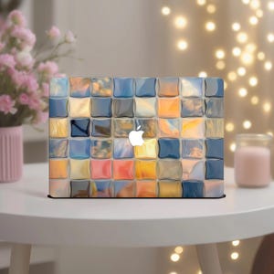 Puede incluir: Un ordenador portátil con un diseño de mosaico abstracto y colorido. El patrón presenta cuadrados en tonos de azul, naranja, amarillo y gris. El logotipo de Apple es visible en el centro. El portátil está sobre una mesa blanca.