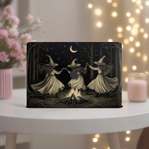 May include: Black laptop skin featuring a sepia-toned illustration of three witches dancing around a bonfire in a forest under a crescent moon. The witches wear pointed hats and flowing dresses. The background includes trees and stars.