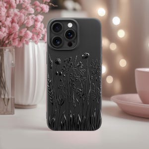 May include: A matte black smartphone case featuring a raised, embossed design of various wildflowers and grasses across the back. The case includes a precise cutout for a triple-lens camera system and a flash, maintaining a sleek, uniform dark finish.