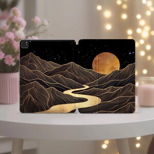 Puede incluir: Una funda para tableta con fondo negro, montañas dibujadas con líneas doradas, un río dorado serpenteante y una gran luna dorada en un cielo estrellado. El diseño se extiende por los paneles frontal y posterior de la cubierta protectora.
