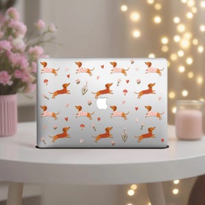 May include: A silver laptop with a repeating pattern of dachshund dogs, hearts, mushrooms, and flowers. Some dogs wear pink polka-dot sweaters. The design is set against a light gray background.