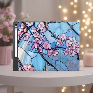 May include: A black tablet case with a floral design featuring pink cherry blossoms and brown branches against a blue stained-glass style background. The tablet is open, revealing the screen and a stylus.