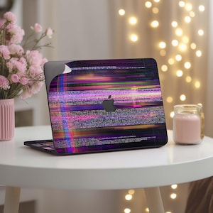 Könnte beinhalten: Ein Laptop mit einem farbenfrohen, abstrakten Design auf dem Deckel. Das Design zeigt horizontale Linien in Lila, Rosa, Grün und Gelb mit einem schwarzen Apple-Logo. Der Laptop ist auf einem weißen Tisch geöffnet, mit einer rosa Kerze und Blumen im Hintergrund.