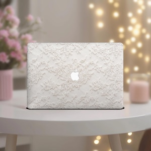 Op de afbeelding: Een witte laptop met een delicaat kanten ontwerp. Het ingewikkelde patroon bedekt het gehele oppervlak, met een prominent Apple-logo in het midden. De laptop staat op een witte tafel, met zachte verlichting en bloemaccenten op de achtergrond.