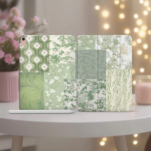 以下が含まれることがあります： 緑と白のパッチワーク模様のiPadケース。花柄、鳥、抽象的な模様が特徴です。ケースは白いテーブルの上に置かれ、背景にはピンクの花瓶とキャンドルがあります。