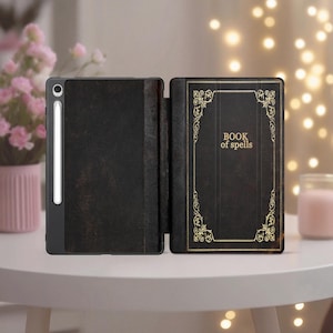 Op de afbeelding: Zwarte tablet hoes met een boekachtig ontwerp. De omslag leest "BOOK of spells" in gouden letters, omlijst door sierlijke gouden krullen. Een stylus is bevestigd. De hoes is open en toont het tabletscherm.