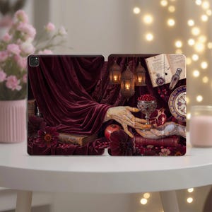 May include: A tablet case featuring a dark red velvet drape, a hand reaching toward a silver goblet filled with pomegranate seeds, hanging lanterns, an open book, a small glass bottle, and dark flowers against a deep, moody background.