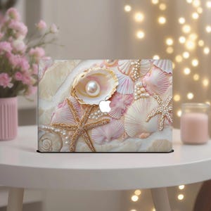 May include: A laptop case featuring a seashell design with a pearl, starfish, and various seashells in shades of pink, white, and gold. The case has an Apple logo and is set against a sandy background.