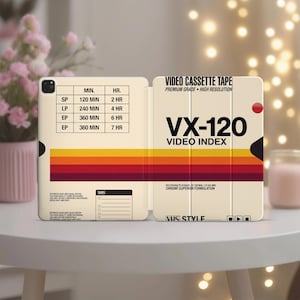 Peut inclure: Une coque de tablette couleur crème arborant un graphisme de cassette VHS rétro. Le design comprend une grille noire avec des durées d'enregistrement, des bandes horizontales jaune, orange et rouge, et le texte VX-120 VIDEO INDEX et VHS STYLE.