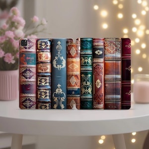 May include: A collection of decorative phone cases designed to resemble antique books. The cases feature intricate designs in various colors, including teal, brown, and burgundy, with gold accents. The cases are arranged upright on a white surface.