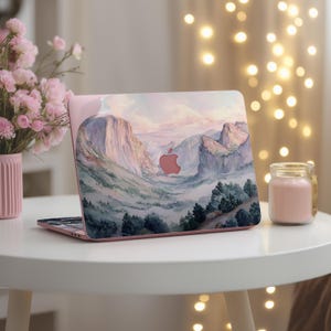 Könnte beinhalten: Ein rosa Laptop mit einem Landschaftsdesign mit Bergen, Bäumen und einem rosa Apple-Logo. Der Laptop ist auf einem weißen Tisch geöffnet, mit einer rosa Blumenvase und einer Kerze im Glas.