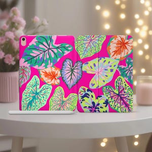 May include: A tablet case featuring a vibrant hot pink background covered in a repeating pattern of stylized heart-shaped leaves in shades of lime green, teal, orange, and purple, accompanied by a slim white stylus resting horizontally in front.