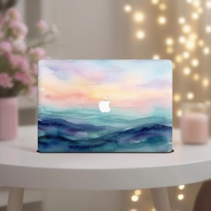 May include: A laptop with a watercolor design featuring a sunset over rolling blue hills. The design has shades of blue, teal, pink, and orange. The Apple logo is visible on the laptop.