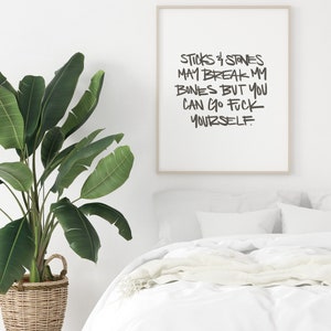 Puede incluir: Un estampado enmarcado con el texto "Sticks & Stones May Break My Bones But You Can Go Fuck Yourself" en letras negras. El estampado est&aacute; en un marco de color claro, exhibido sobre una cama con ropa de cama blanca. Una gran planta en maceta est&aacute; en primer plano.