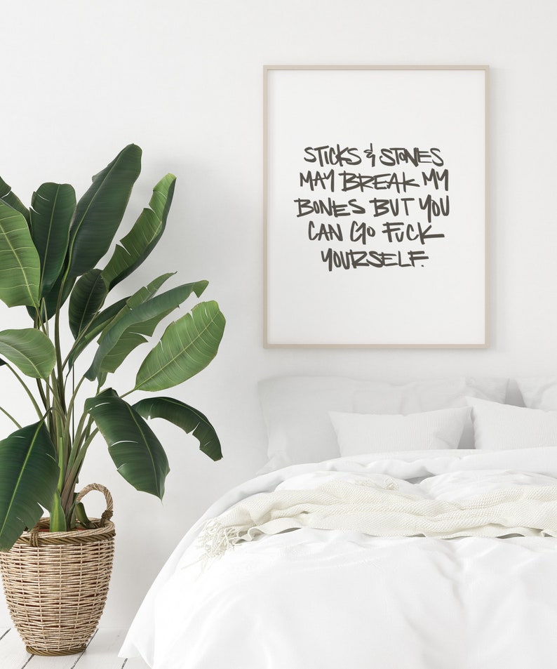 Puede incluir: Un estampado enmarcado con el texto "Sticks & Stones May Break My Bones But You Can Go Fuck Yourself" en letras negras. El estampado est&aacute; en un marco de color claro, exhibido sobre una cama con ropa de cama blanca. Una gran planta en maceta est&aacute; en primer plano.