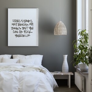 Puede incluir: Un marco con el texto "STICKS & STONES MAY BREAK MY BONES BUT YOU CAN GO FUCK YOURSELF" cuelga sobre una cama con ropa de cama blanca. Tambi&eacute;n se ven una l&aacute;mpara colgante tejida y un jarr&oacute;n alto.