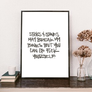 Puede incluir: Una impresi&oacute;n enmarcada con el texto "Sticks & Stones May Break My Bones But You Can Go F*** Yourself" en un estilo manuscrito. La impresi&oacute;n se muestra sobre una superficie de madera oscura con libros, una taza blanca y flores secas en jarrones de vidrio.