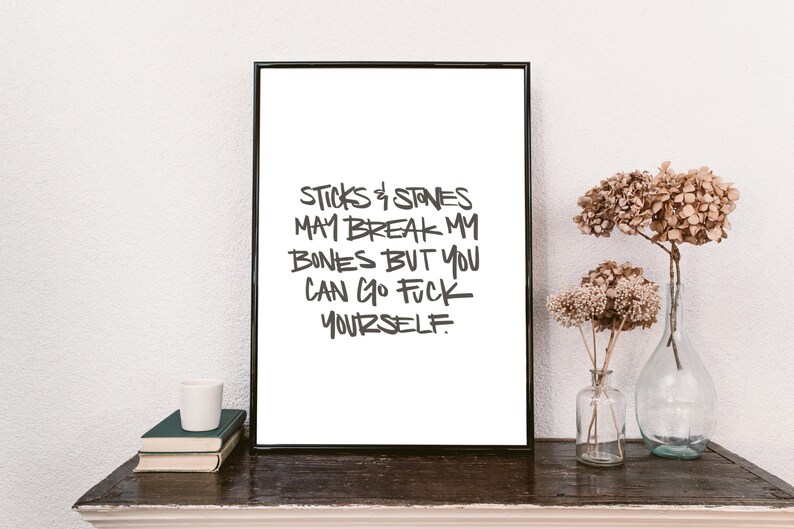 Puede incluir: Una impresi&oacute;n enmarcada con el texto "Sticks & Stones May Break My Bones But You Can Go F*** Yourself" en un estilo manuscrito. La impresi&oacute;n se muestra sobre una superficie de madera oscura con libros, una taza blanca y flores secas en jarrones de vidrio.