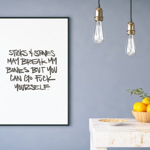 Puede incluir: Impresi&oacute;n enmarcada en blanco con texto negro que dice: "Sticks & Stones May Break My Bones But You Can Go Fuck Yourself". La impresi&oacute;n cuelga de una pared azul sobre una mesa blanca con un cuenco de madera con naranjas.
