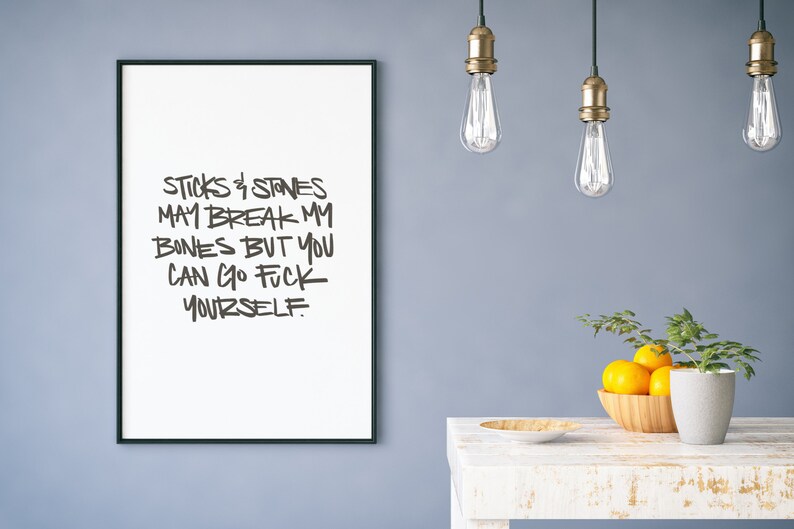 Puede incluir: Impresi&oacute;n enmarcada en blanco con texto negro que dice: "Sticks & Stones May Break My Bones But You Can Go Fuck Yourself". La impresi&oacute;n cuelga de una pared azul sobre una mesa blanca con un cuenco de madera con naranjas.
