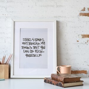 Puede incluir: Un marco blanco con el texto "STICKS & STONES MAY BREAK MY BONES BUT YOU CAN GO FUCK YOURSELF." El marco est&aacute; sobre un escritorio blanco con una maceta, l&aacute;pices, libros y una taza.