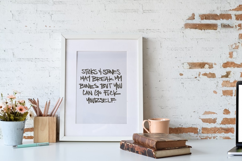 Puede incluir: Un marco blanco con el texto "STICKS & STONES MAY BREAK MY BONES BUT YOU CAN GO FUCK YOURSELF." El marco est&aacute; sobre un escritorio blanco con una maceta, l&aacute;pices, libros y una taza.