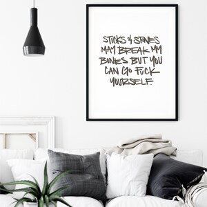 Puede incluir: Un cuadro enmarcado con el texto "STICKS & STONES MAY BREAK MY BONES BUT YOU CAN GO FUCK YOURSELF" en un marco negro. El cuadro cuelga sobre un sof&aacute; blanco con cojines grises y blancos. Una l&aacute;mpara colgante negra cuelga del techo.