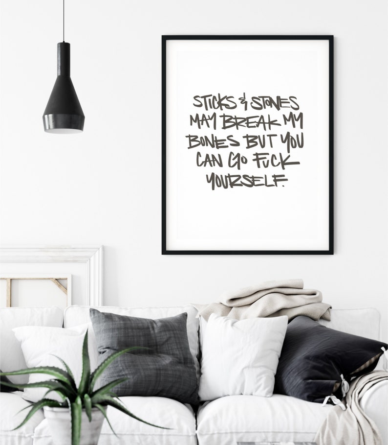 Puede incluir: Un cuadro enmarcado con el texto "STICKS & STONES MAY BREAK MY BONES BUT YOU CAN GO FUCK YOURSELF" en un marco negro. El cuadro cuelga sobre un sof&aacute; blanco con cojines grises y blancos. Una l&aacute;mpara colgante negra cuelga del techo.