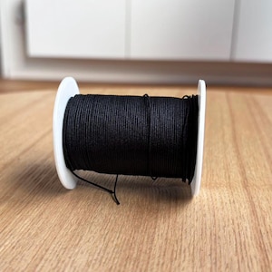May include: A spool of black elastic cord wound around a white plastic bobbin.