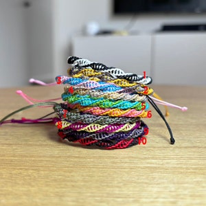 Könnte beinhalten: Ein Stapel von zehn bunten Freundschaftsarmbändern aus Fäden in verschiedenen Farben. Die Armbänder sind übereinander gestapelt, wobei das oberste Armband schwarz-weiß ist.