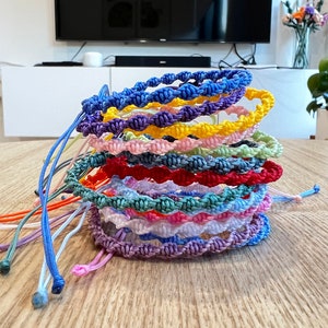 May include: A stack of colourful braided bracelets in various shades of blue, purple, pink, yellow, green, and red. The bracelets are made of woven string and have a wavy pattern.