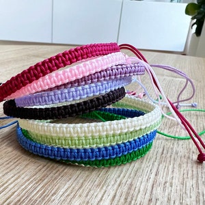 May include: A stack of eight colourful braided bracelets in various shades of pink, purple, brown, white, green, and blue. The bracelets are made of woven cord and have adjustable ties.