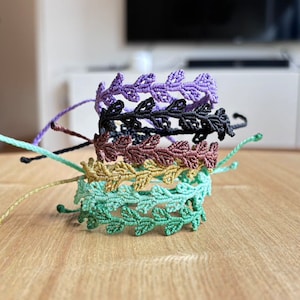 Könnte beinhalten: Ein Stapel von vier handgefertigten Freundschaftsarmbändern mit einem Blattdesign. Die Armbänder sind in den Farben Grün, Gold, Braun und Lila.