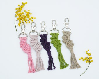 May include: Five colorful macrame keychains with silver key rings. The keychains are made with a spiral pattern and have tassels at the bottom. The colors are pink, beige, purple, green, and natural.