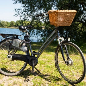 May include: A grey bicycle with a wicker basket on the handlebars, a black seat, and black mudguards. The bicycle is parked on a grassy path near a lake.