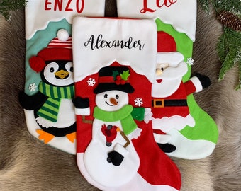 May include: Three Christmas stockings with personalized names: Enzo, Alexander, and Leo. The stockings feature festive designs, including a penguin, snowman, and Santa Claus. The stockings are red, green, and white with decorative accents.