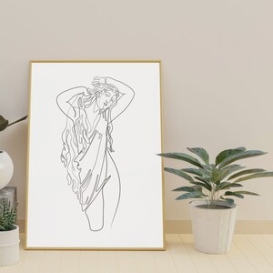 May include: A black and white line drawing of a person with long hair, wearing a flowing garment, with their arms raised above their head. The drawing is framed in a gold frame and is hanging on a wall.