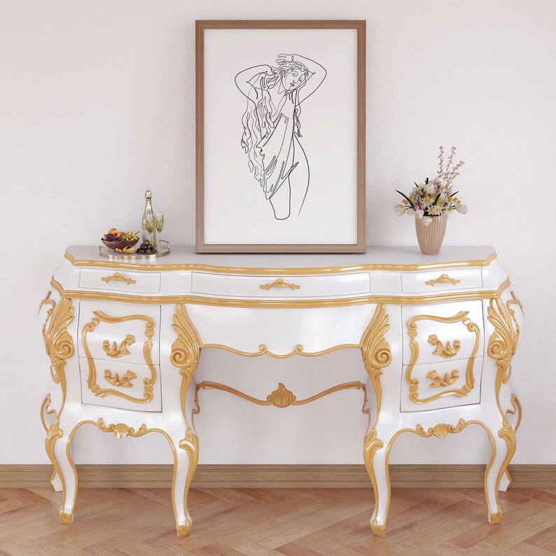 May include: White and gold ornate vanity table with three drawers on each side. The table has a curved top and ornate legs. A framed line drawing of a woman is hanging on the wall above the table.