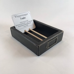May include: A rectangular, black wooden business card holder with a distressed finish. The holder features two wooden dowels and a metal label holder. A business card with the text "Whippersnapper Creates" is displayed.
