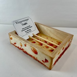 May include: A wooden business card holder with a hinged lid. The holder is decorated with a fall leaf design in shades of red and orange. The business card inside reads "Whippersnapper Creates". Two wooden dowels are inside the holder.