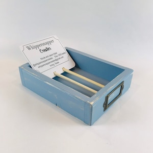 May include: A rectangular, light blue wooden box with a distressed finish. The box has a small metal label holder and holds two light-colored dowels. A business card with the text "Whippersnapper Creates" is inside the box.