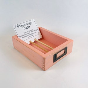 May include: A rectangular, light pink wooden business card holder with a distressed finish. It features a metal label holder and two wooden dowels. A business card with the text "Whippersnapper Creates" is displayed.