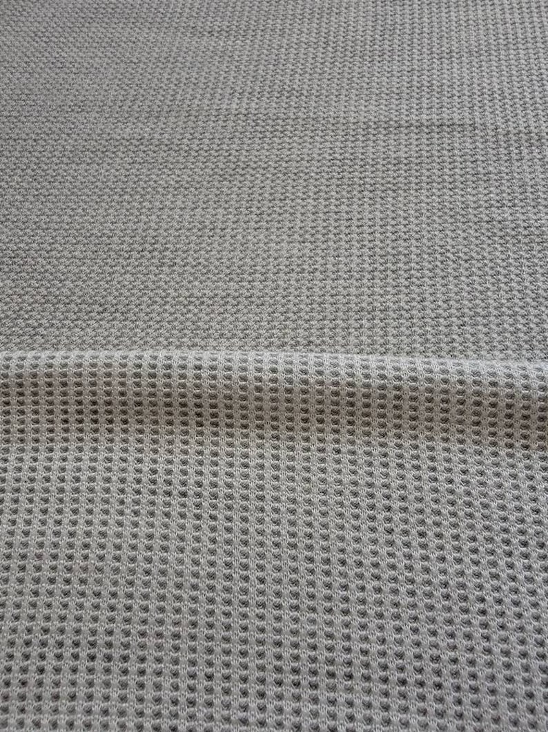 May include: A close-up of a gray waffle knit fabric. The fabric has a textured surface and a subtle sheen.